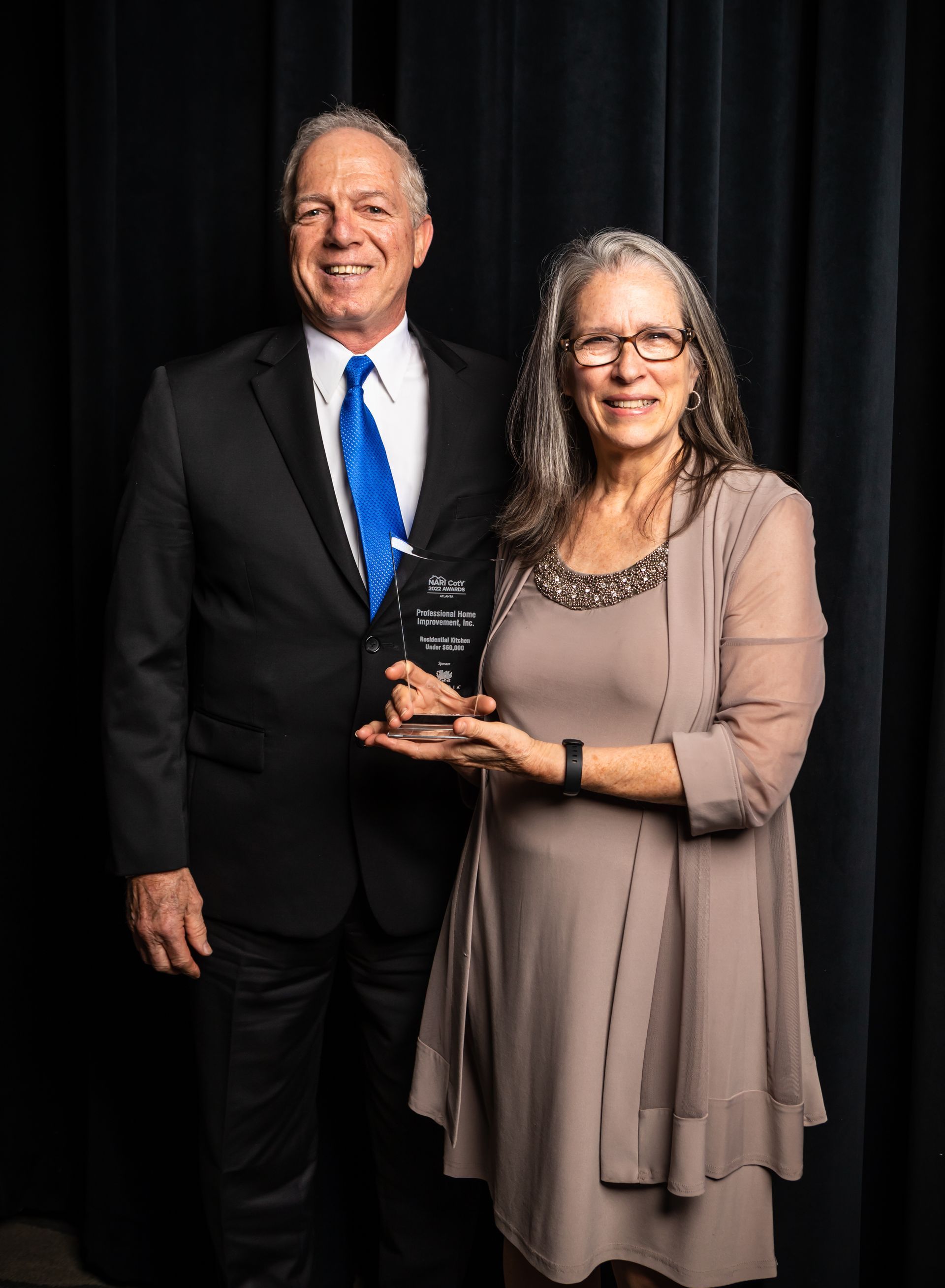 Two people in formal attire holding a clear award trophy in front of a black curtain.
