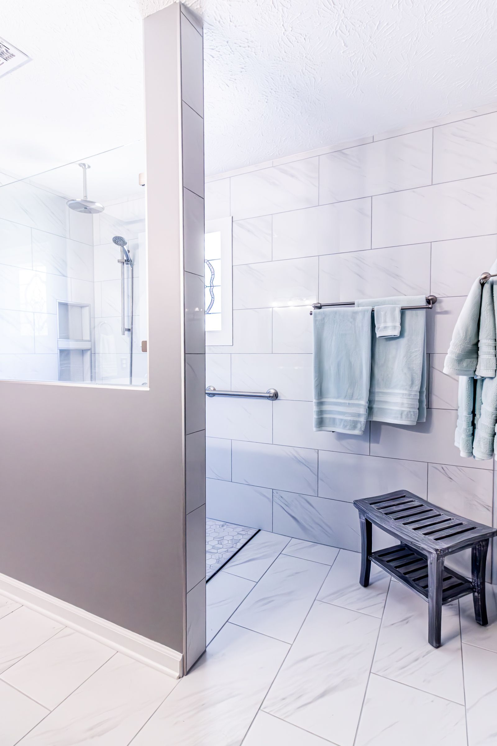 Modern tiled bathroom with glass shower, wall-mounted towels, and a black bench