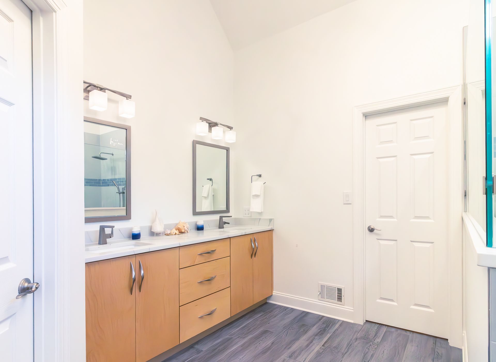 Bright bathroom vanity with mirrors, white walls, wood cabinets, and a tiled floor near a closed door.