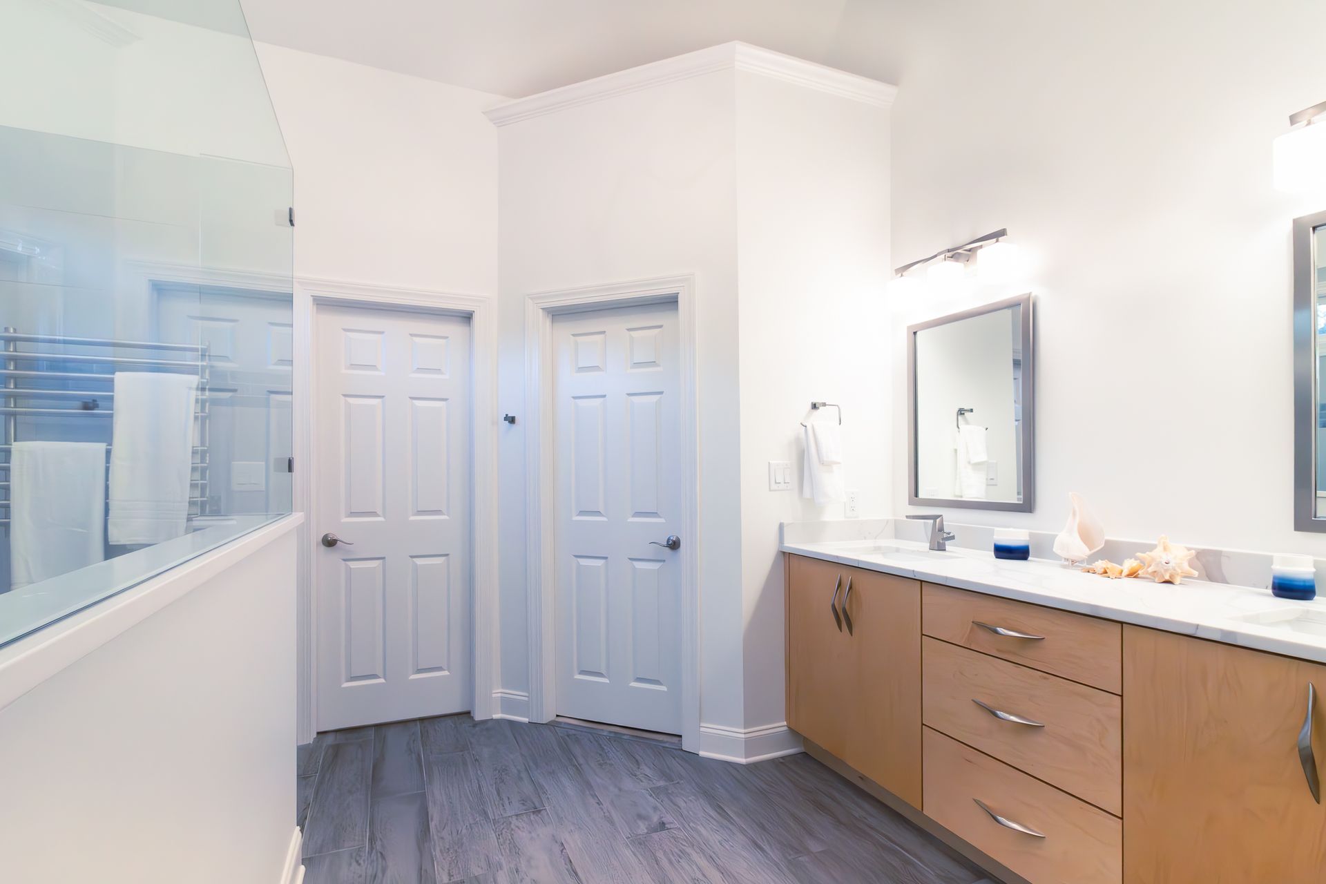 Bright bathroom with white walls, double doors, wood vanity, mirror, and gray flooring