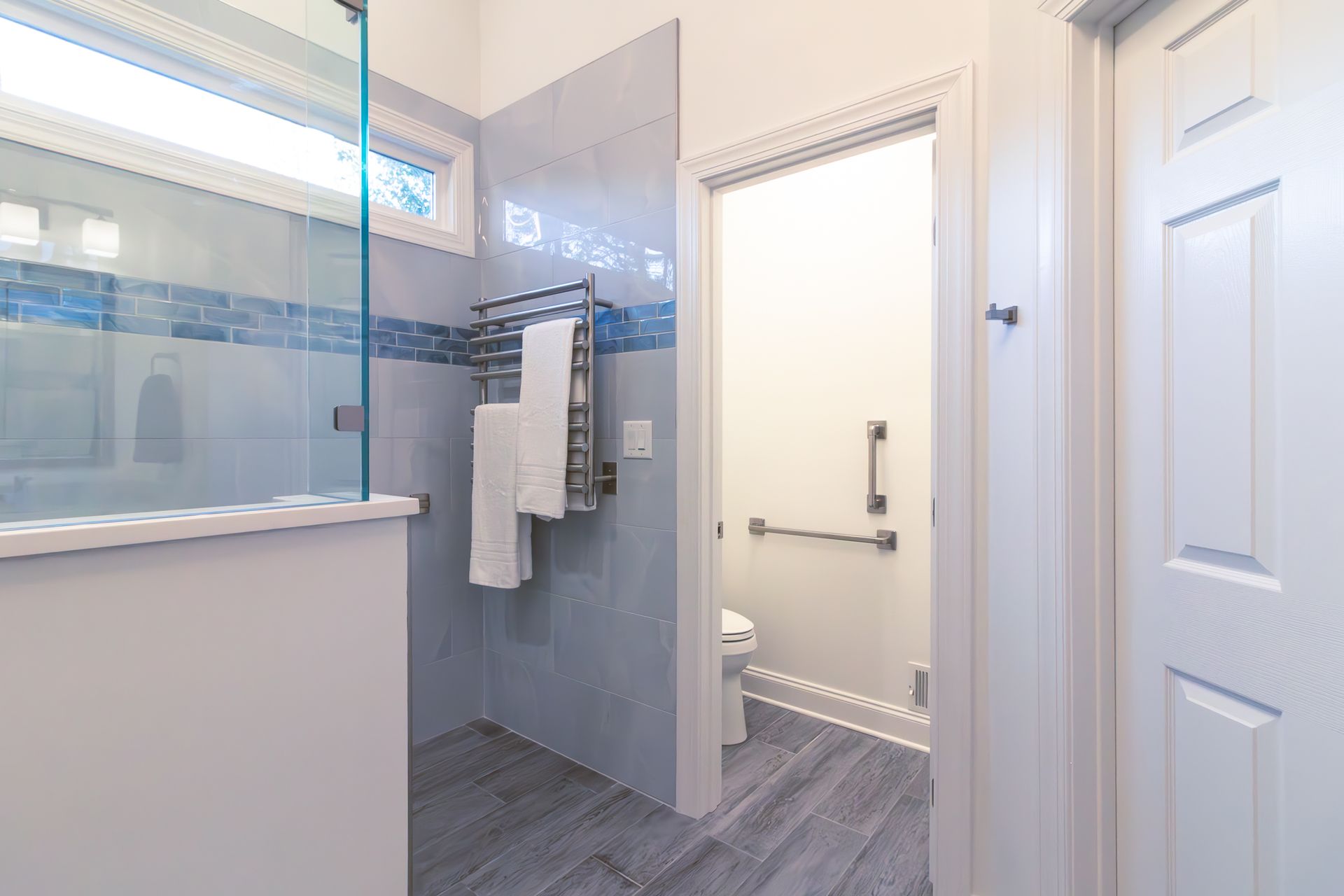 Modern bathroom with glass shower, towel rack, and open doorway to a bright adjoining room