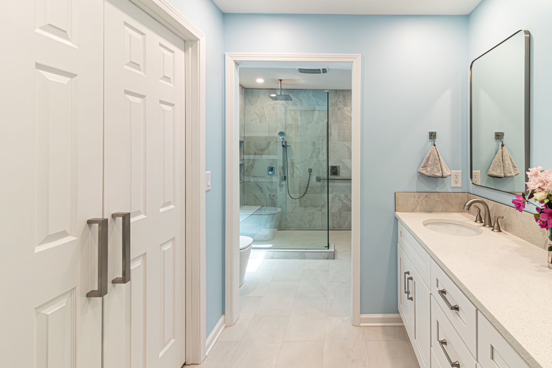 Bright bathroom vanity leading to a glass-enclosed shower with white cabinets and light blue walls