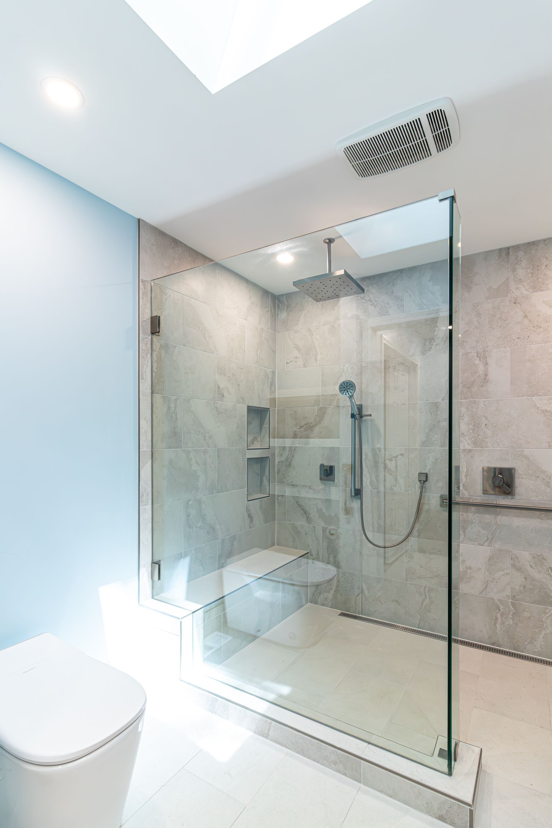 Modern walk-in shower with glass enclosure, marble walls, rain showerhead, and toilet in a bright bathroom