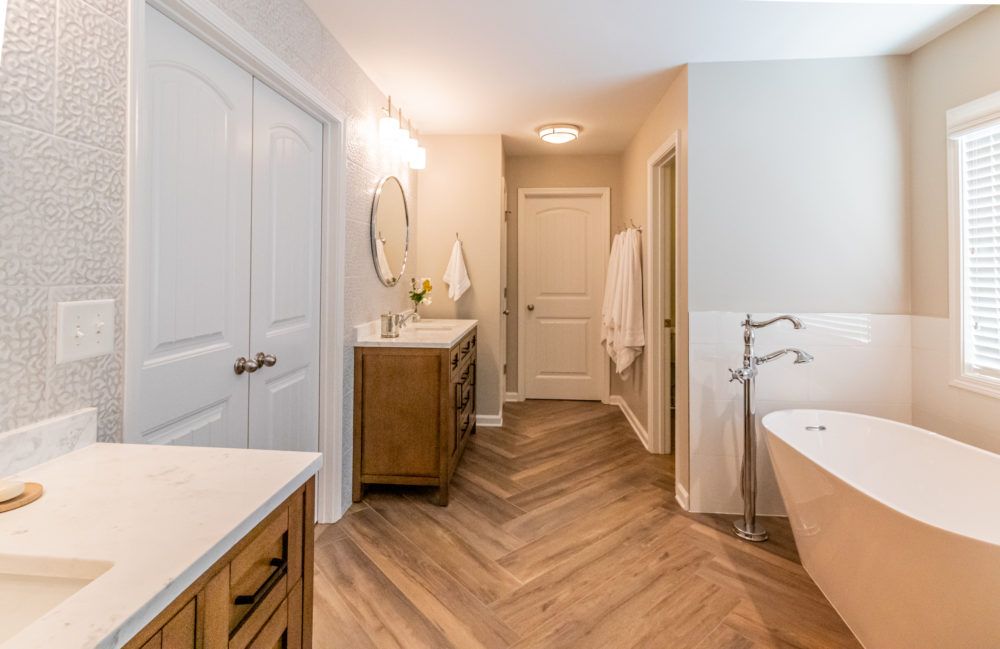 Bright bathroom with double vanity, freestanding tub, herringbone floor, and light neutral decor.