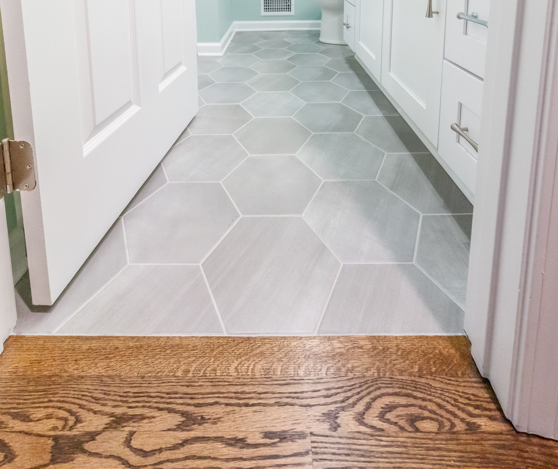 Hallway with light gray hex tile floor leading from a wood floor threshold into a white bathroom
