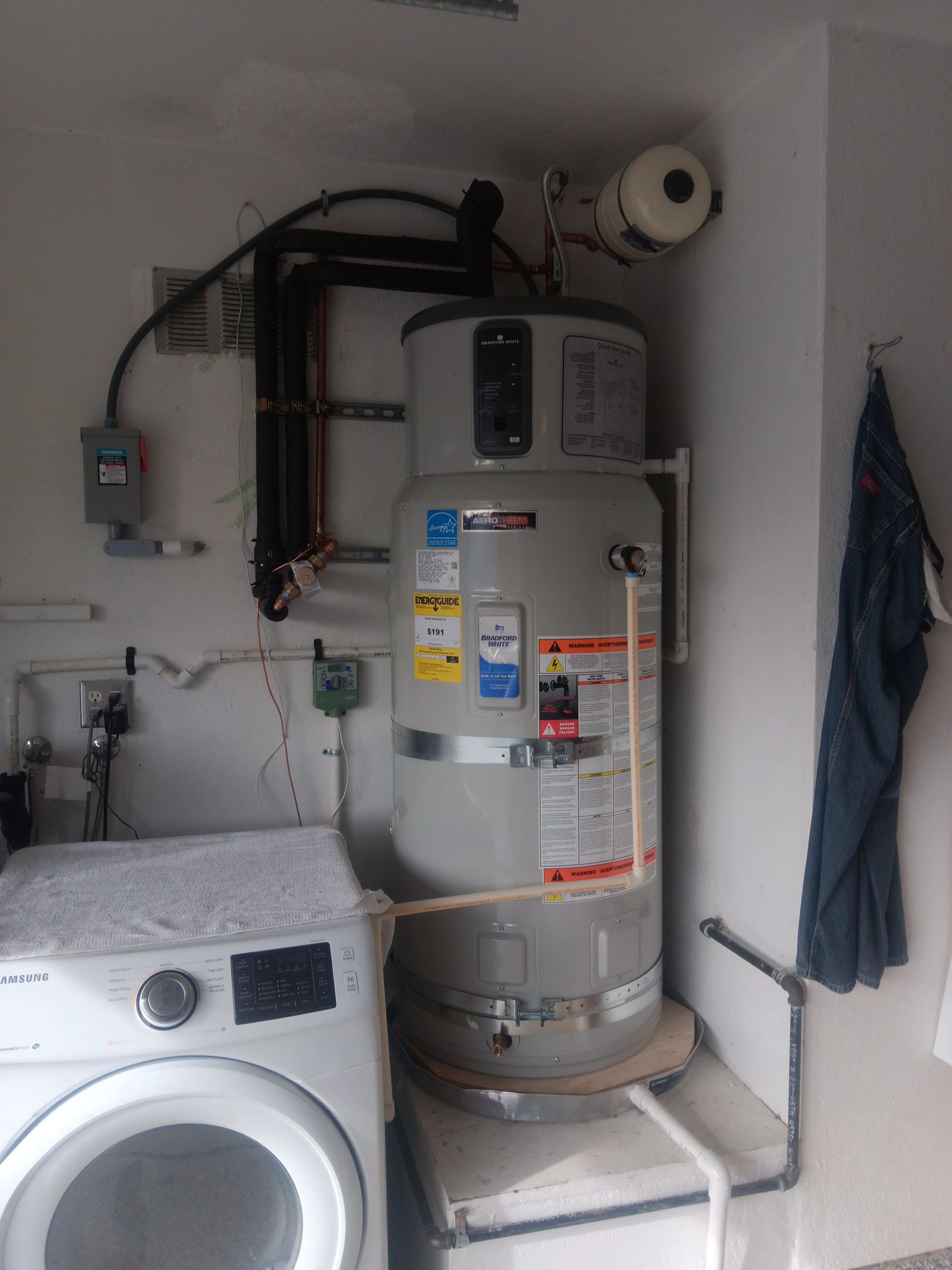 Gray water heater in a narrow closet with exposed pipes and warning labels.
