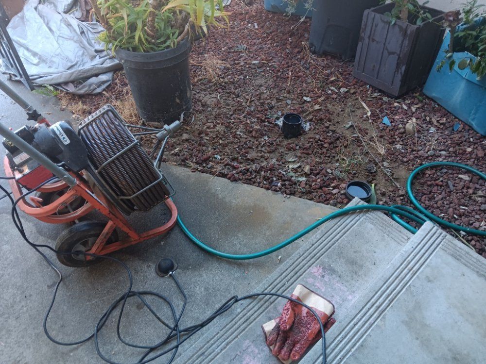 Garden corner with a red hose reel, green hose, potted plants, and mulch beside a concrete patio.