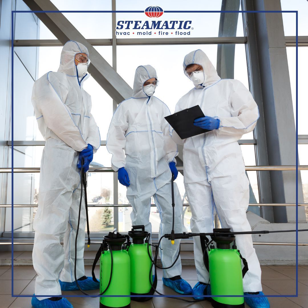 Three workers in white protective suits inspect green tanks in a industrial facility.
