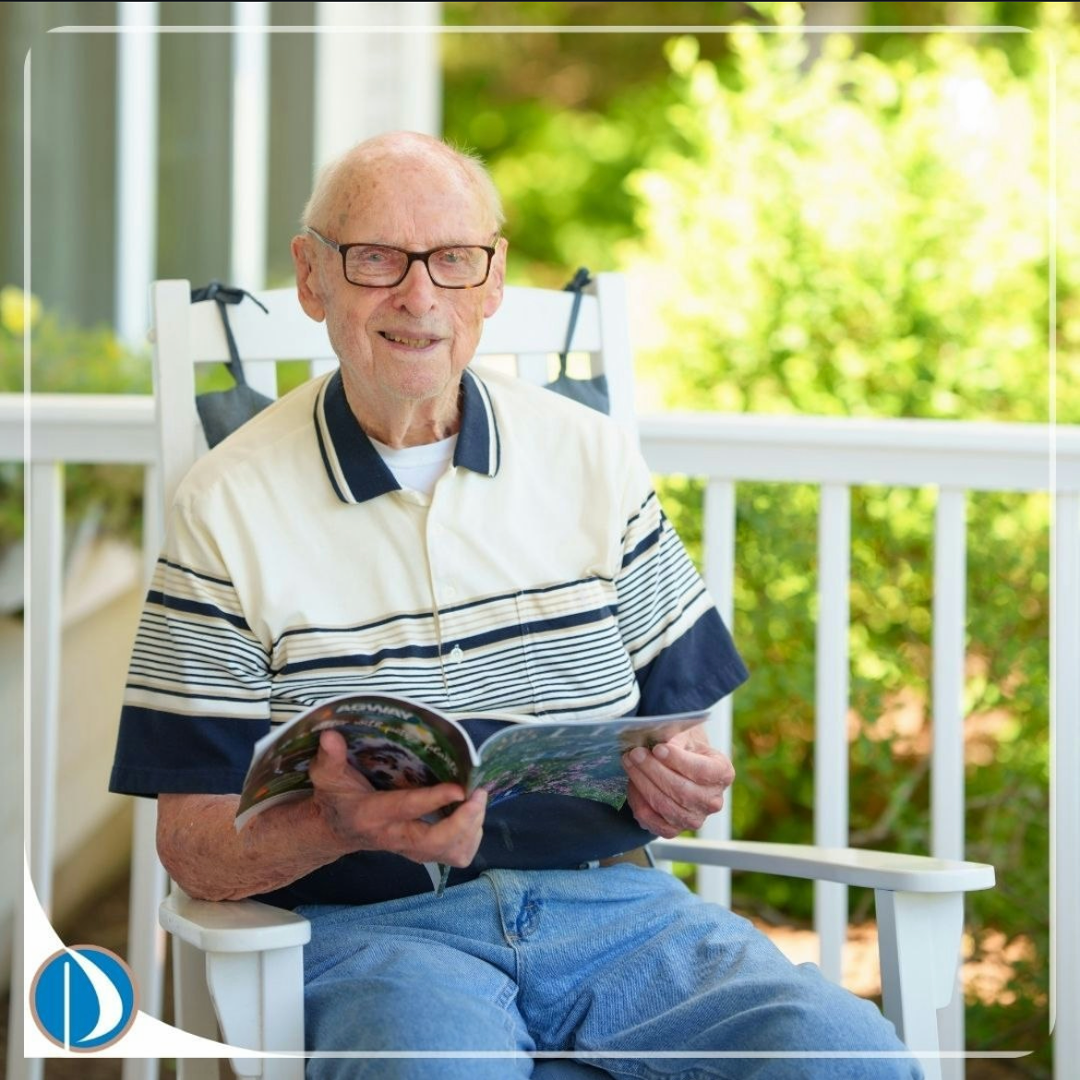 Older man in glasses reading a magazine on a white porch chair beside a railing and greenery.