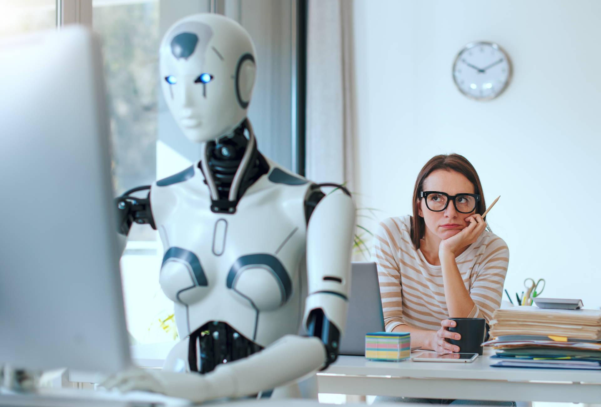 A woman looking uncertain at her desk while a humanoid robot works on a computer beside her