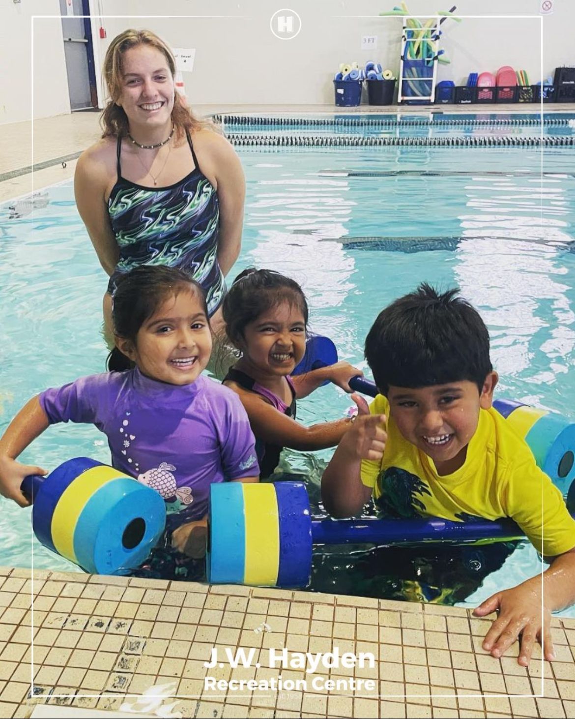 Children with pool noodles smiling in an indoor swimming pool, with a woman standing behind them.