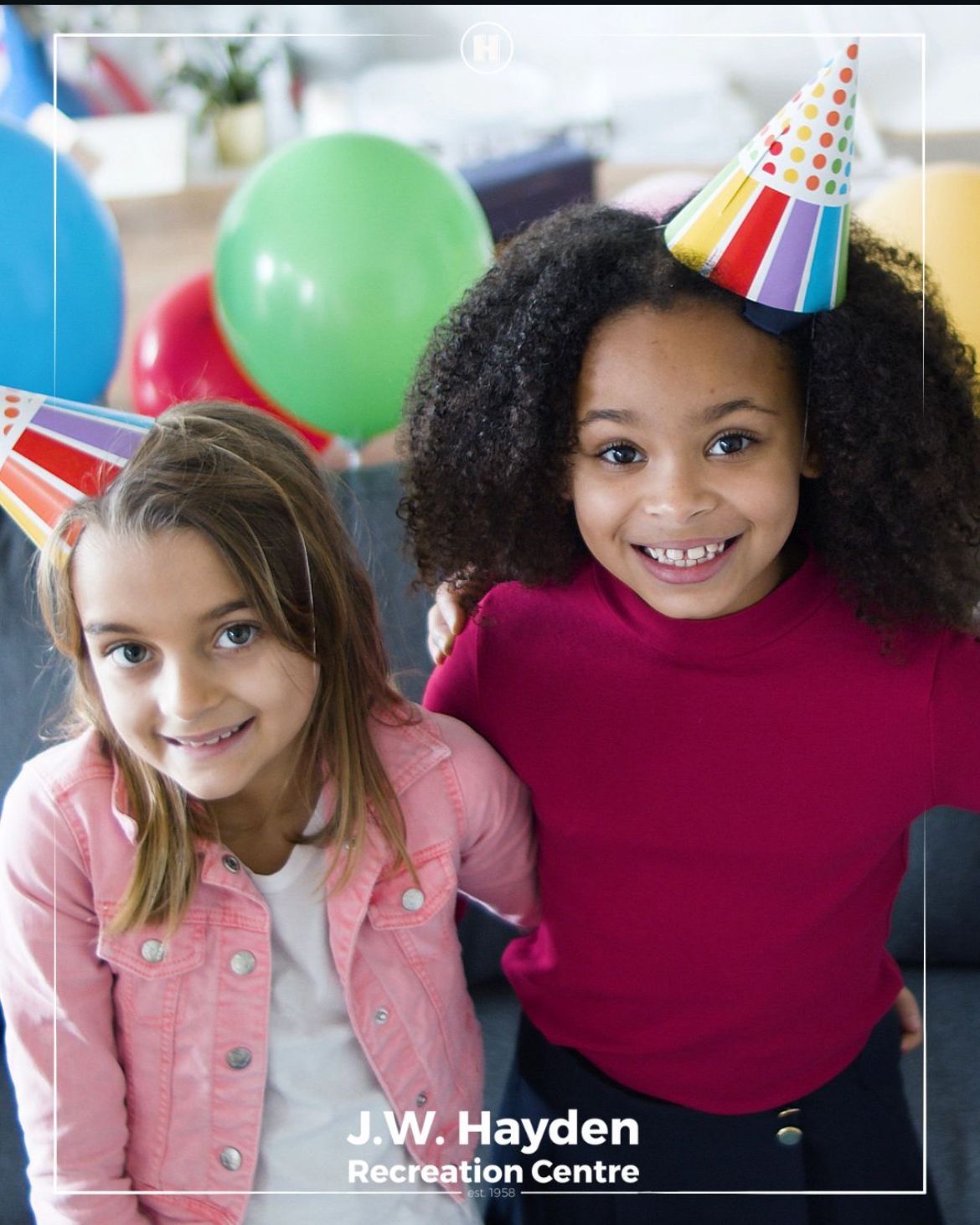 Two smiling children wearing party hats with balloons in the background