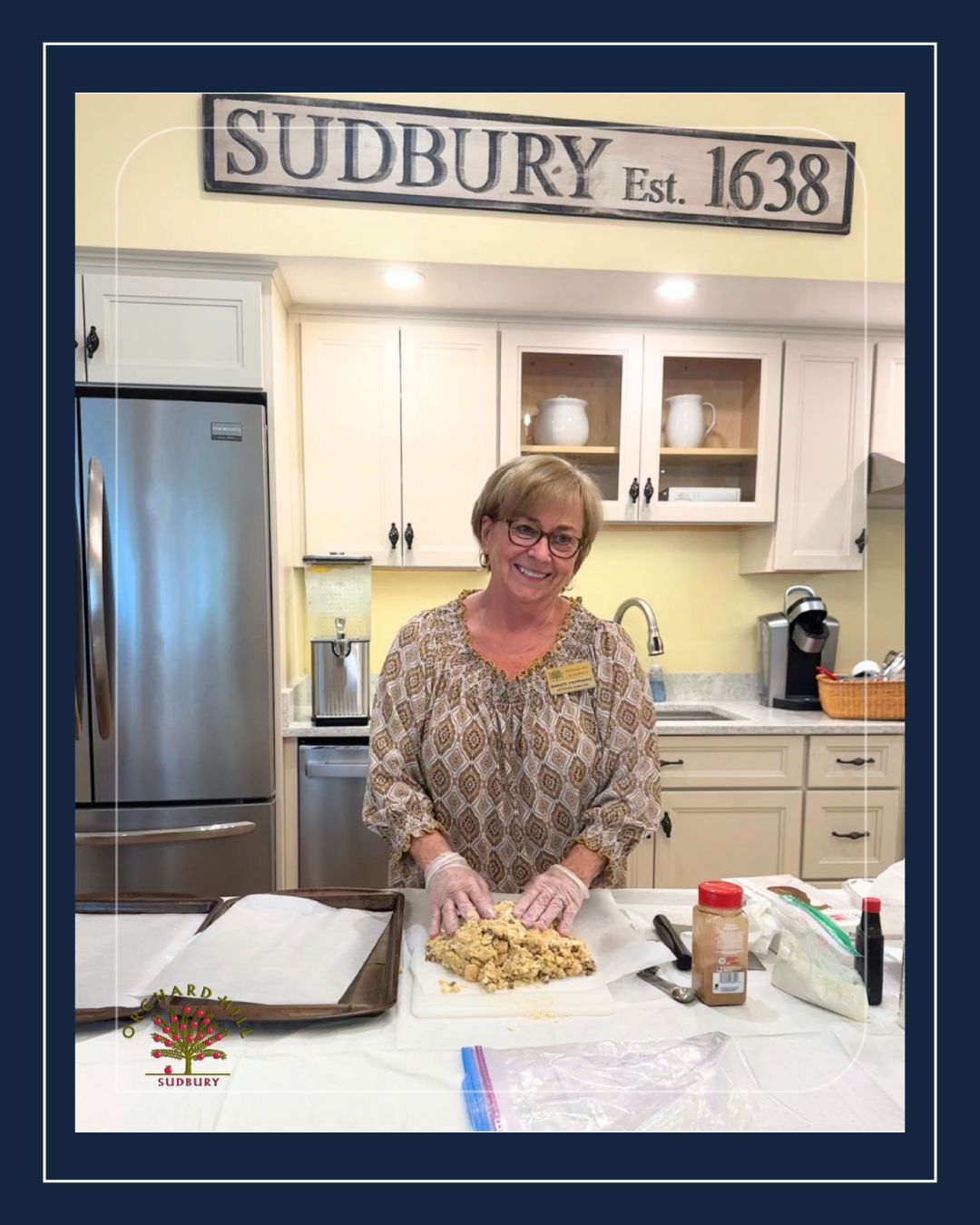 Woman mixing dough in a bright kitchen with a “SUDBURY Est. 1638” sign above the cabinets