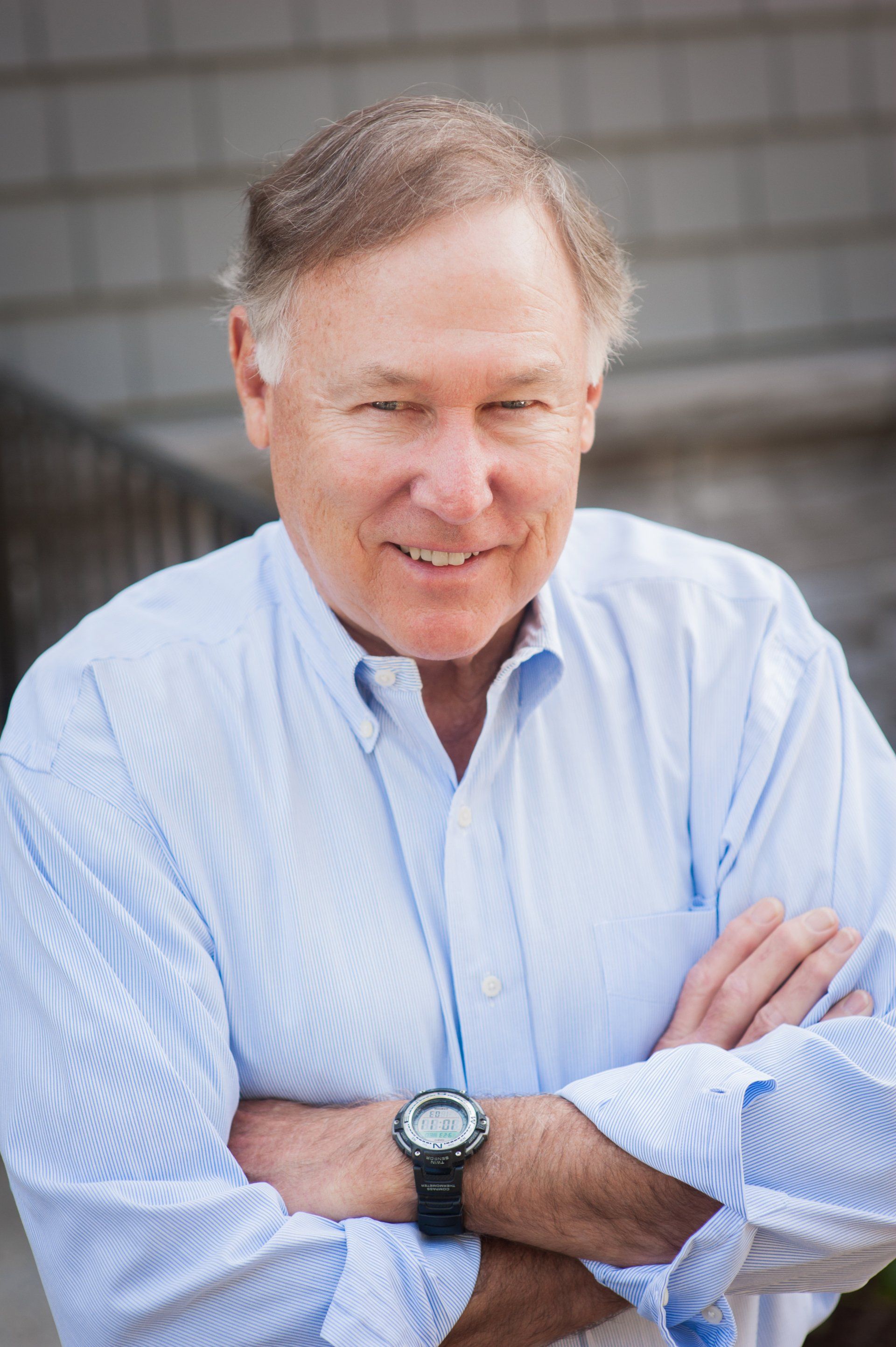 Smiling man in a light blue shirt with arms crossed, standing outdoors against a blurred background