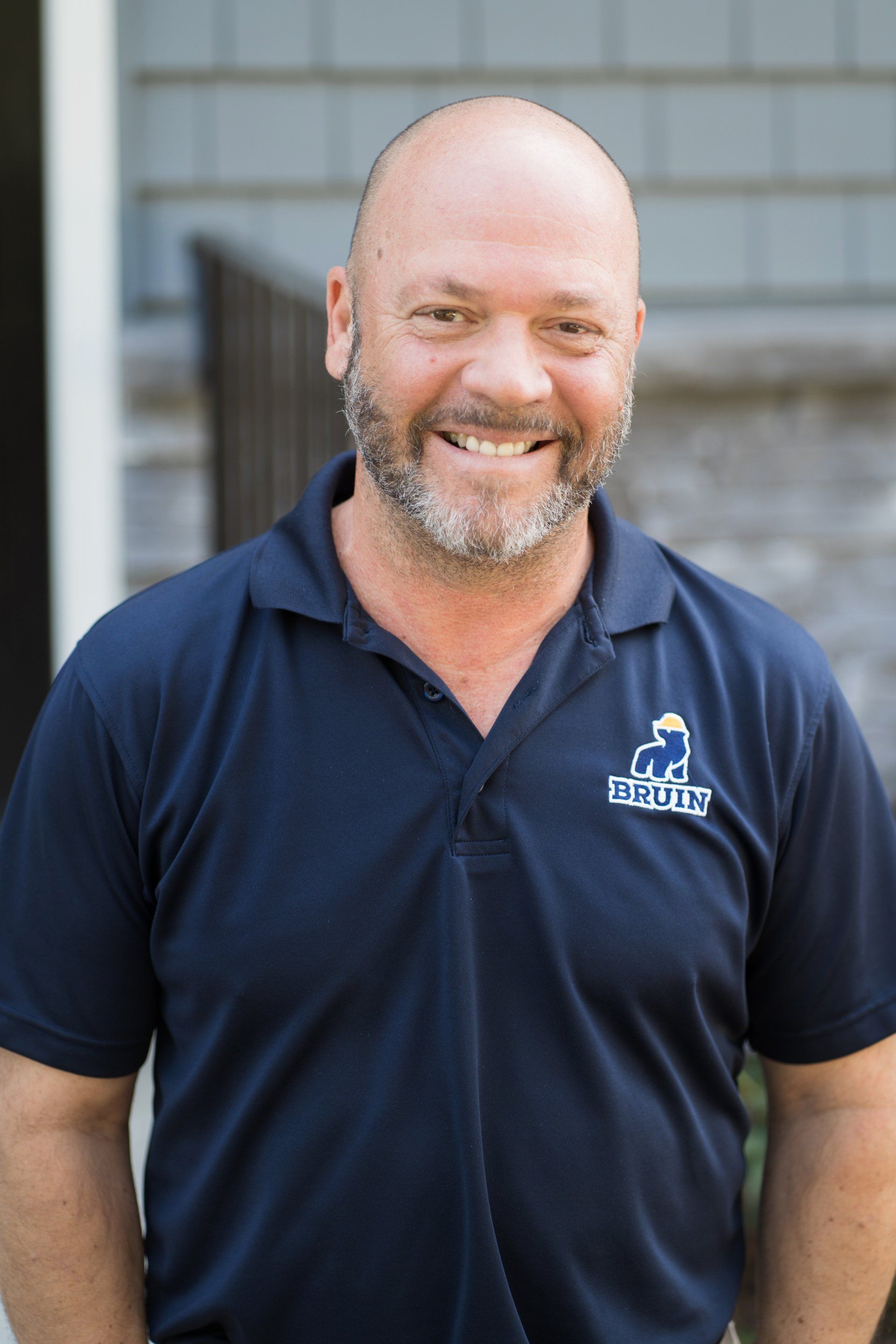 Smiling bald man in a navy polo shirt standing outdoors against a blurred building background