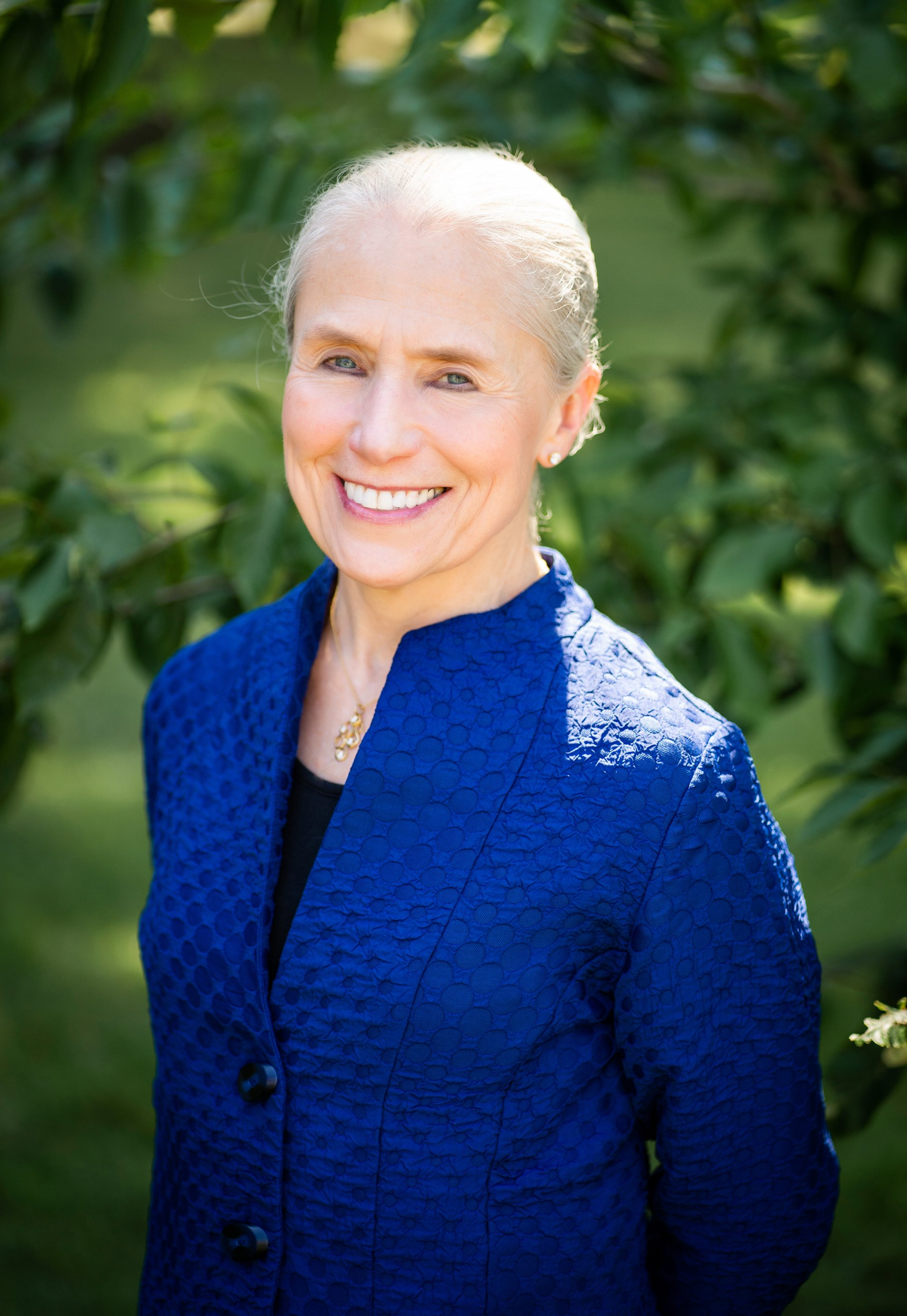 Smiling person in a cobalt blue jacket standing outdoors against leafy green foliage