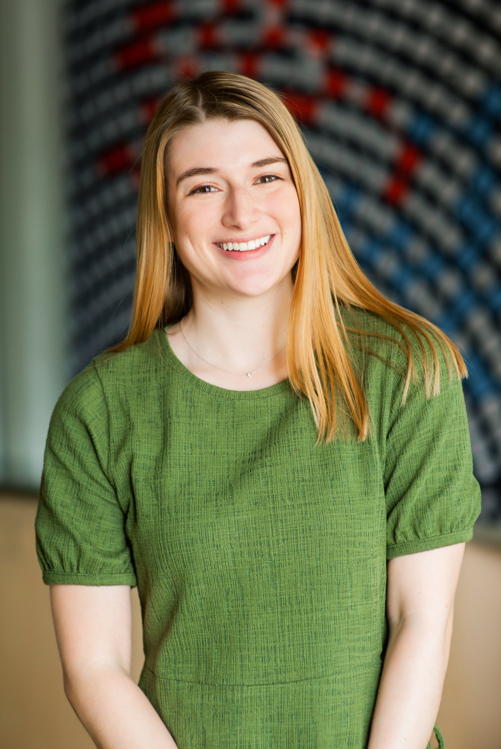 Smiling person in a green shirt standing indoors before a blurred patterned backdrop