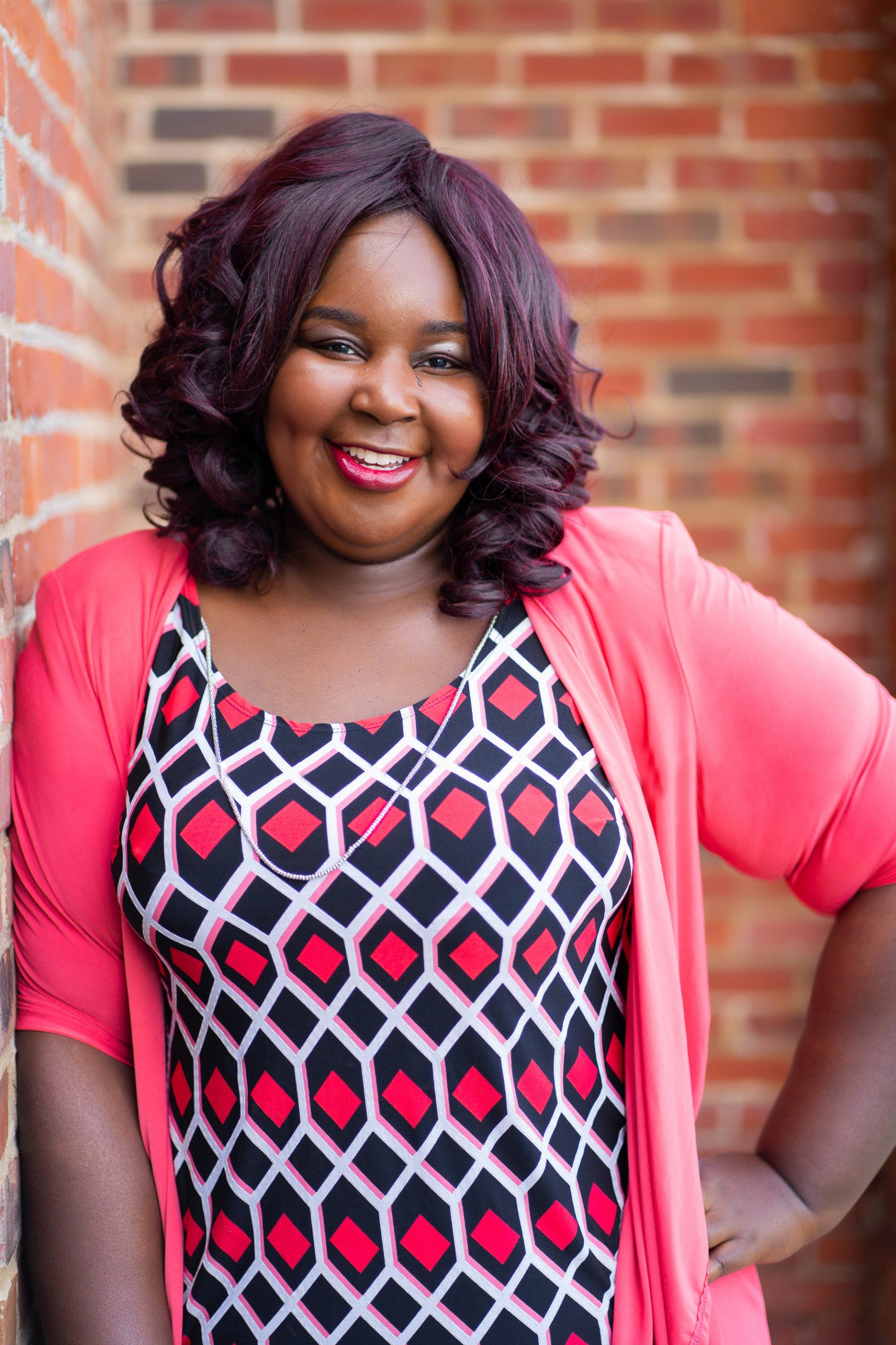 Smiling person in a pink cardigan and patterned dress standing before a brick wall