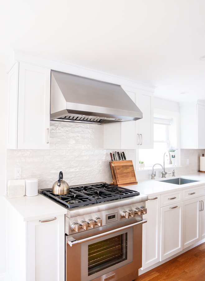 Bright white kitchen with stainless steel stove, hood, and white cabinets under a window