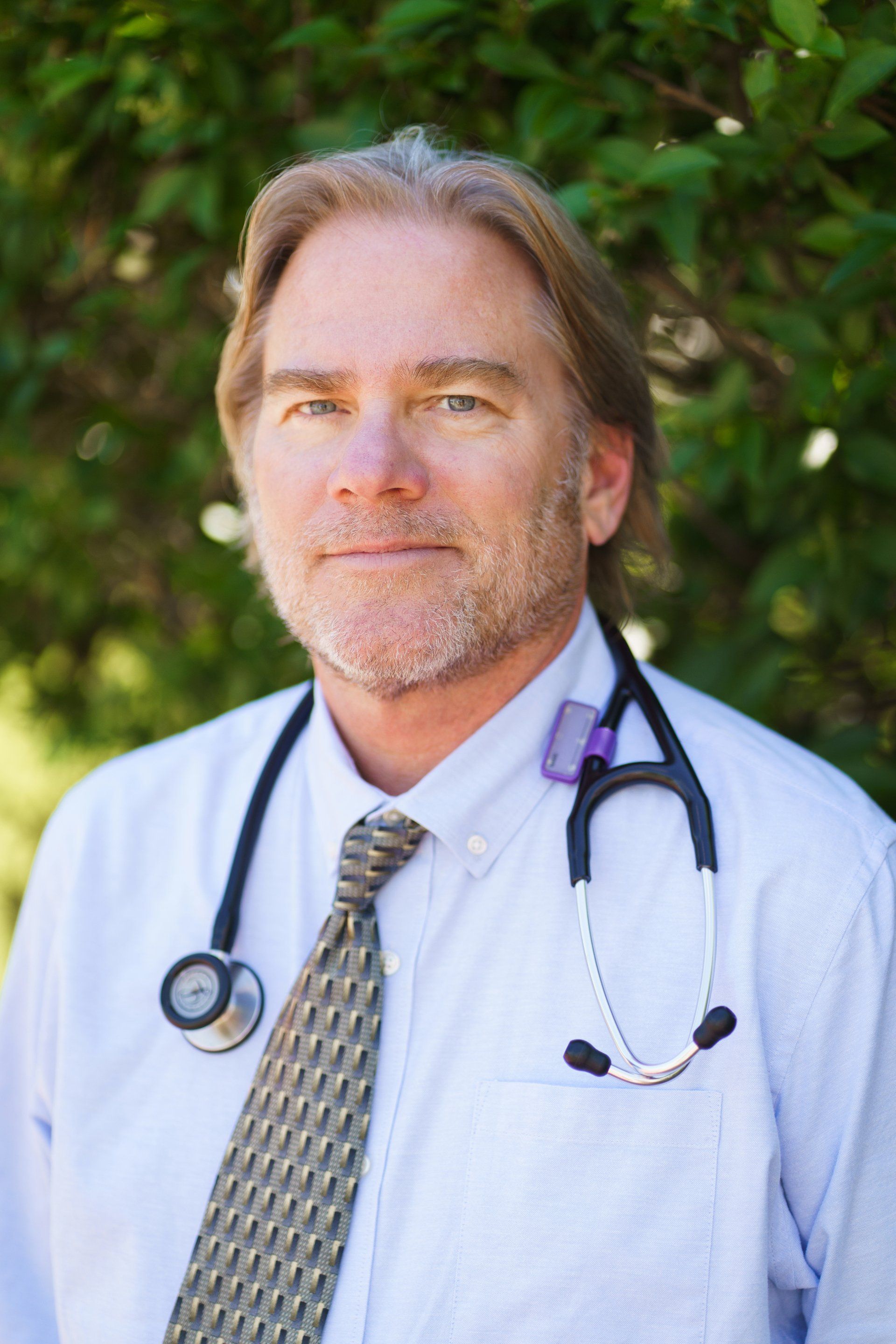 Doctor in a white coat with a stethoscope standing outdoors against greenery