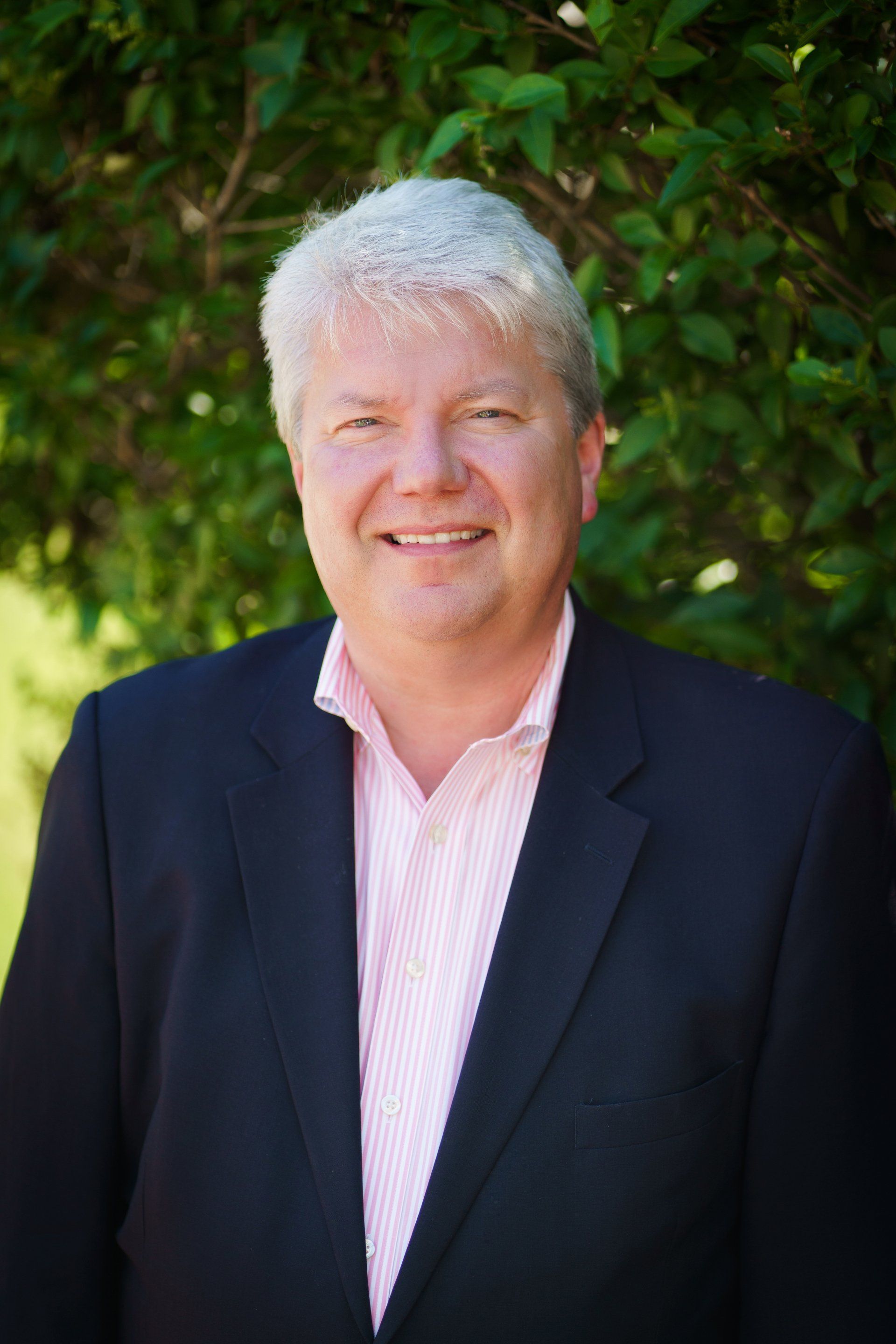 Smiling man in a dark blazer and pink shirt standing outdoors in front of green foliage