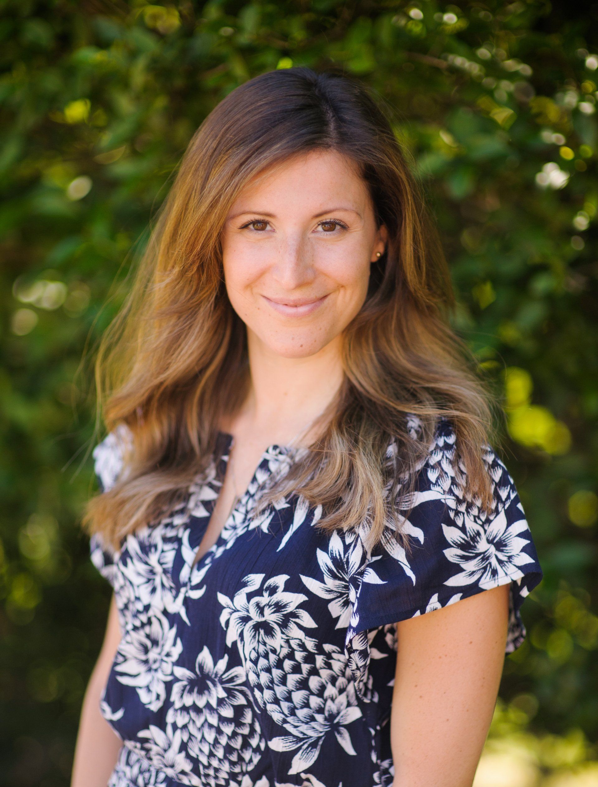 Smiling woman in a navy floral dress standing outdoors against a green leafy background