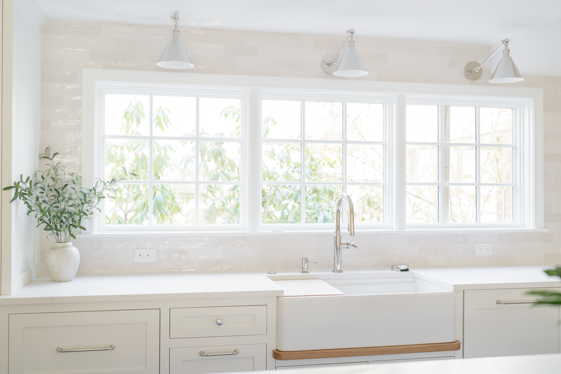 Bright white kitchen sink under large sunlit windows with a potted plant and pendant lights