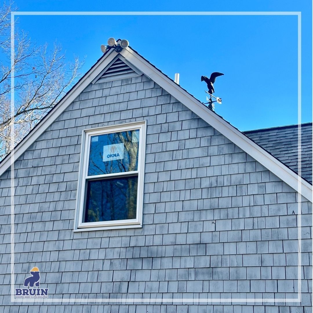 Gray house gable with a small window against a bright blue sky; a small figure stands on the roof ridge.