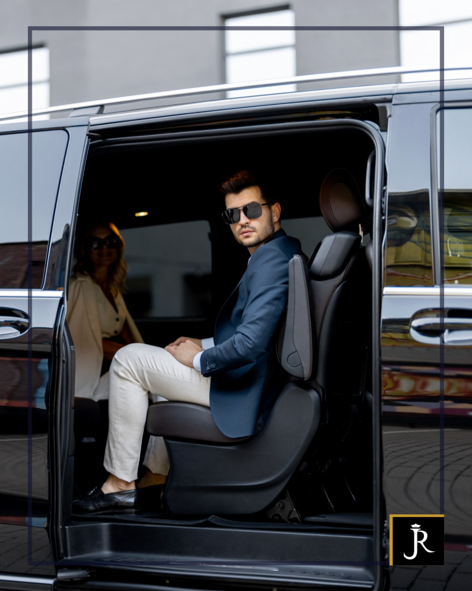 Man in sunglasses sitting in a black van doorway, wearing a blue blazer and beige pants.