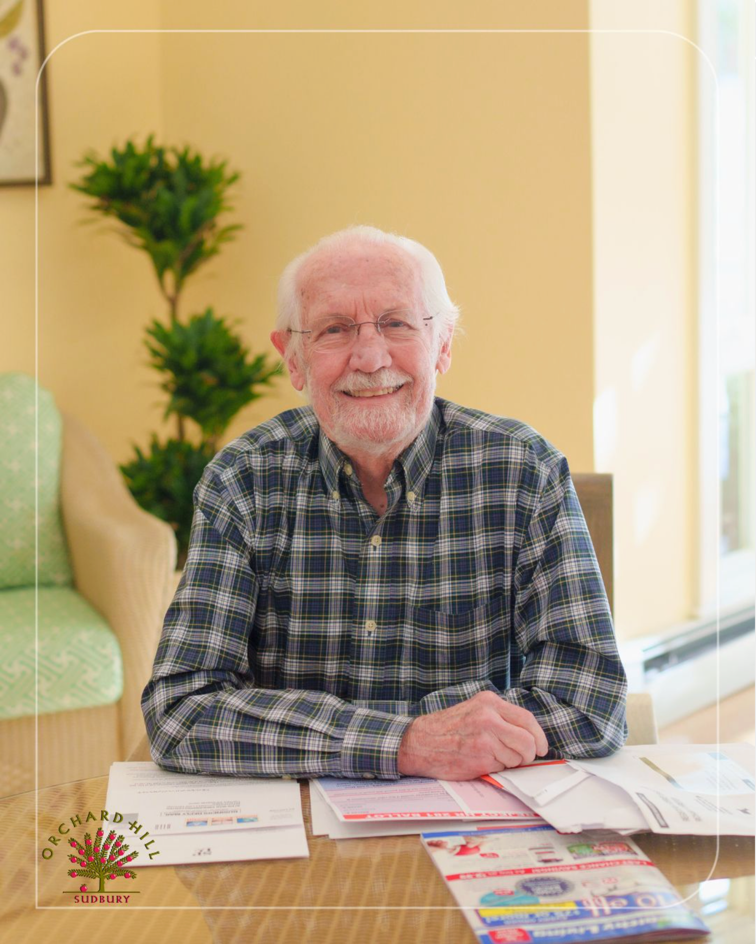 Smiling man in plaid shirt seated at a table with brochures and papers in a bright room