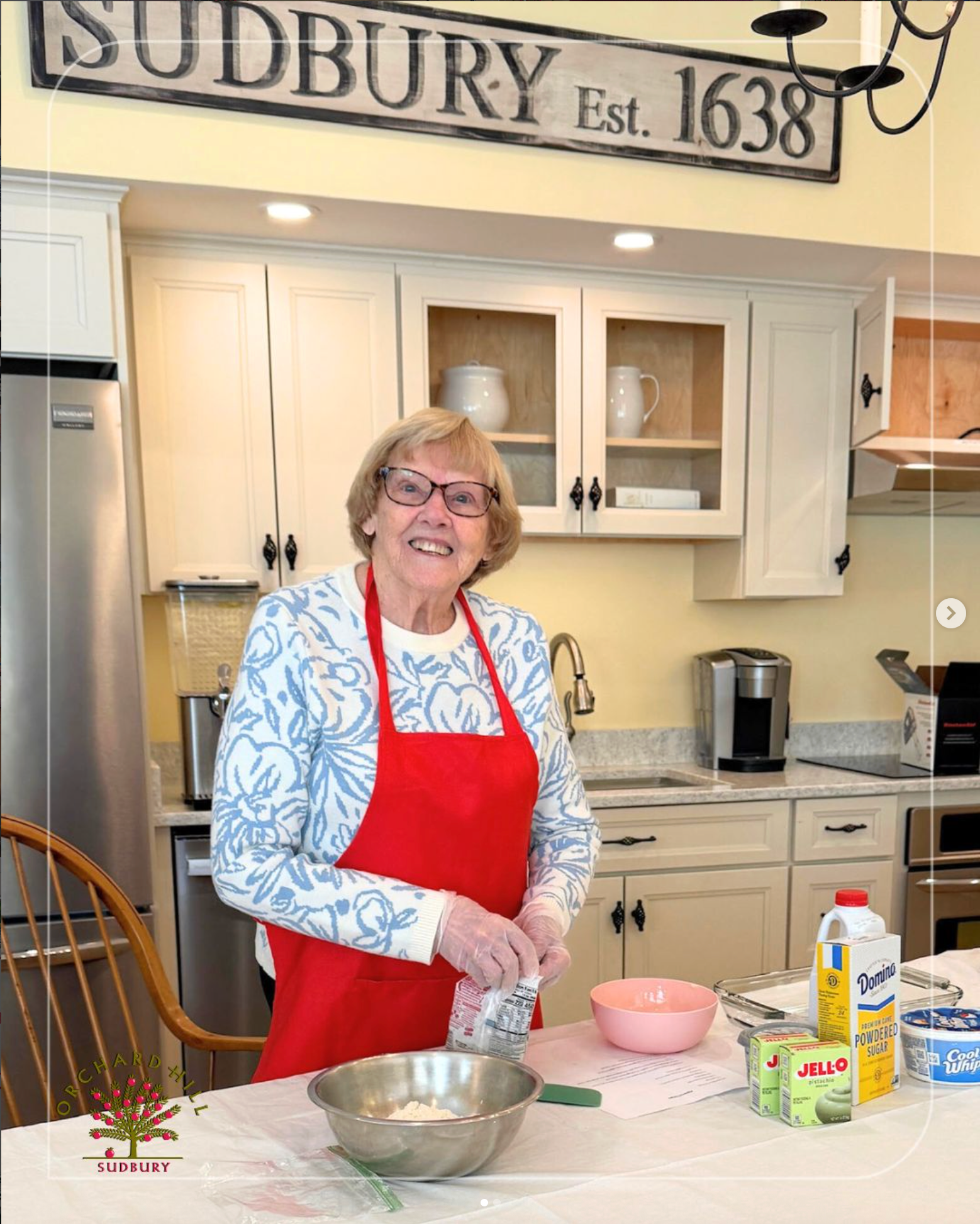 Woman in red apron mixing ingredients in a kitchen under a “Sudbury Est. 1638” sign