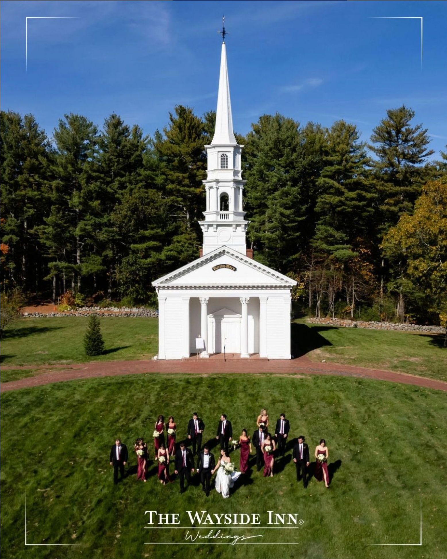 White church with tall steeple on a lawn, with a small group standing in front, under a blue sky.
