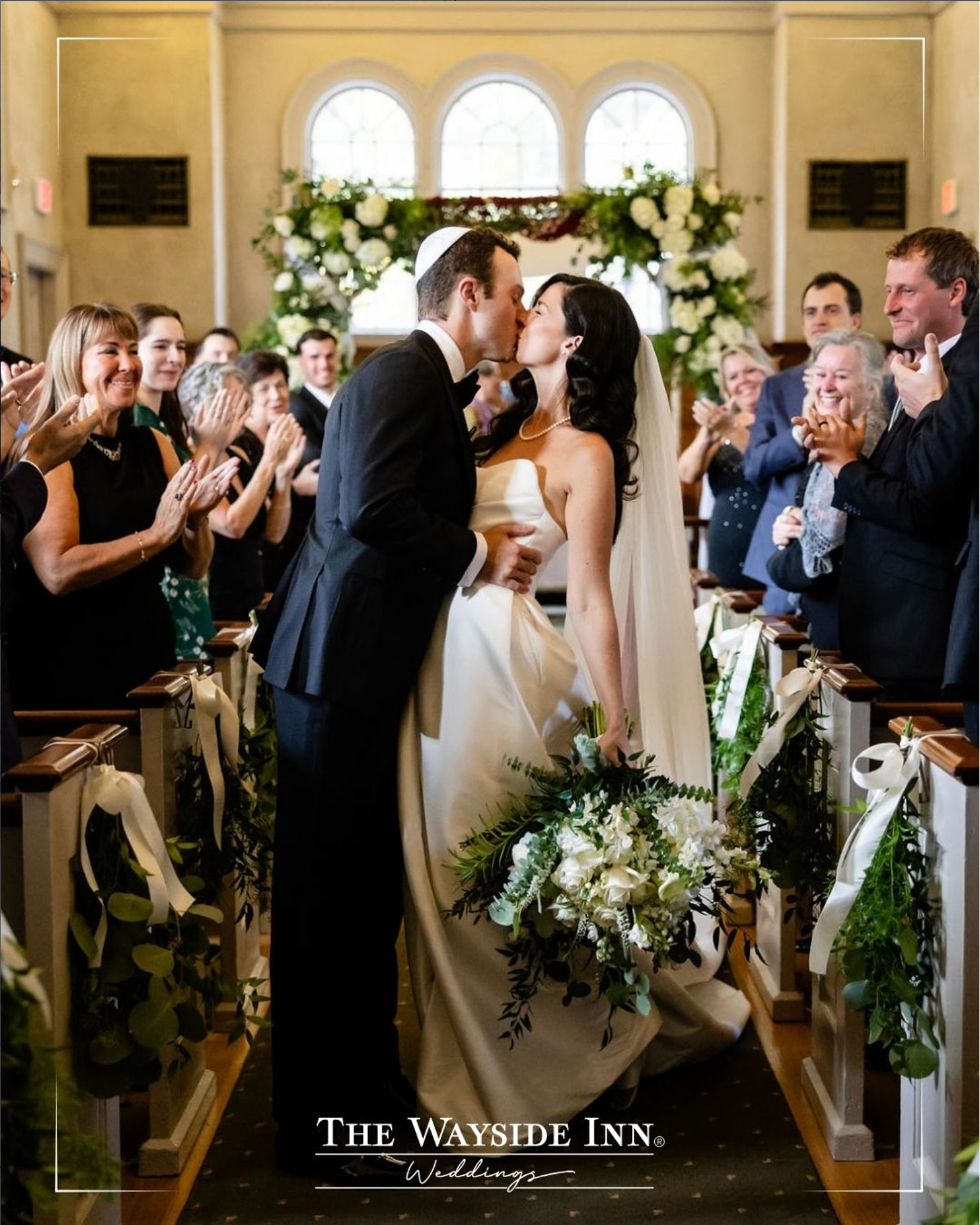 Wedding couple kissing at the altar in a flower-lined church aisle, guests clapping on both sides