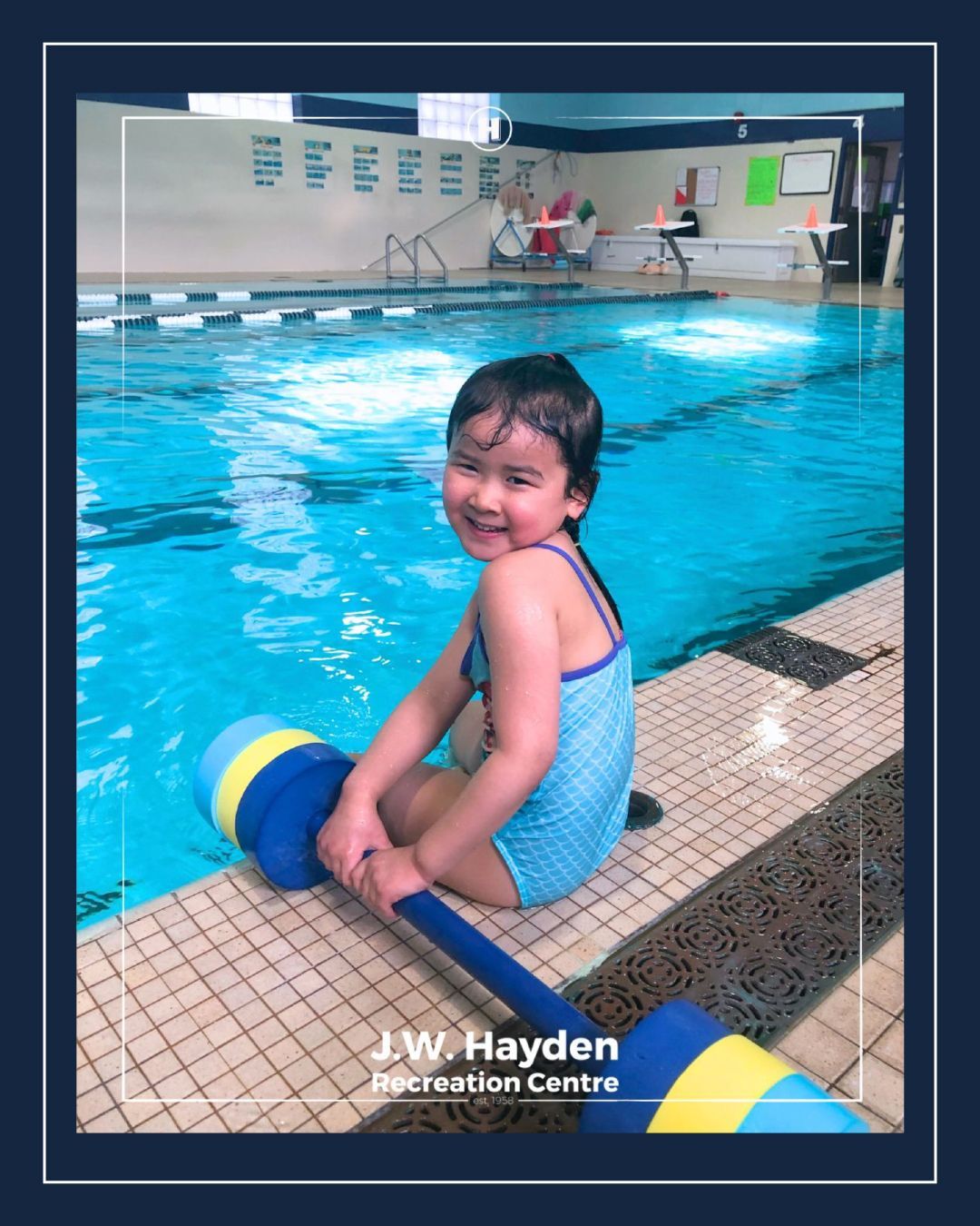 Child smiling by an indoor pool, sitting on a blue-and-yellow foam float, with JW Hayden Recreation Centre in background