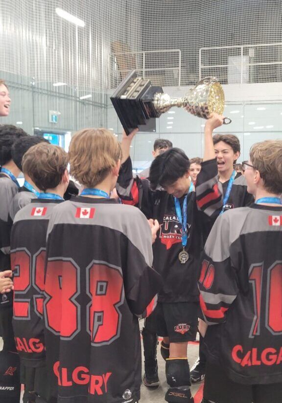 Fury Ball Hockey team in black jerseys gathers around a player holding up a gold trophy at the BHA Provincials