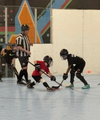Two floor hockey players in black and red crouch for a faceoff as a referee watches.