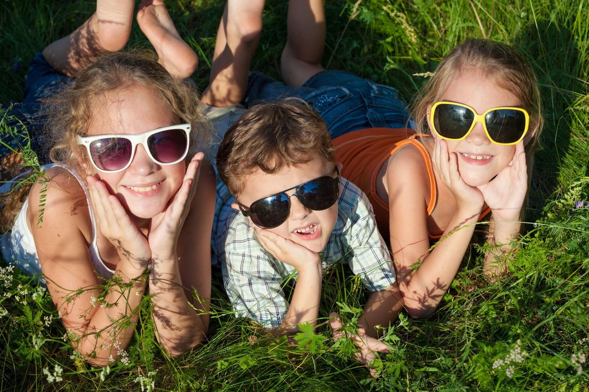 Tre bambini sdraiati sull'erba, sorridenti con gli occhiali da sole, con i piedi di un altro bambino visibili dietro di loro.