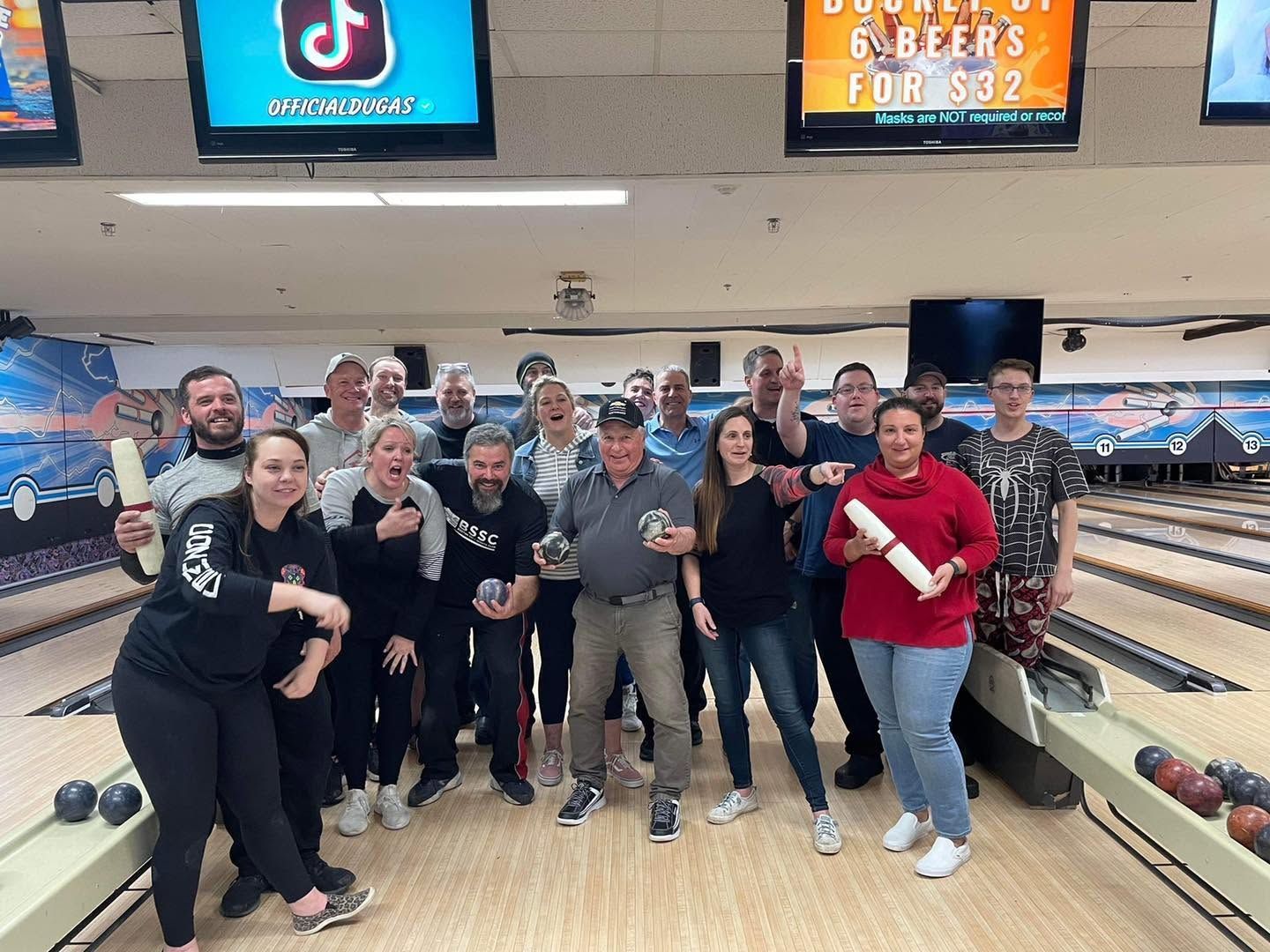Group of people posing with bowling balls and pins in a bowling alley, smiling and celebrating.