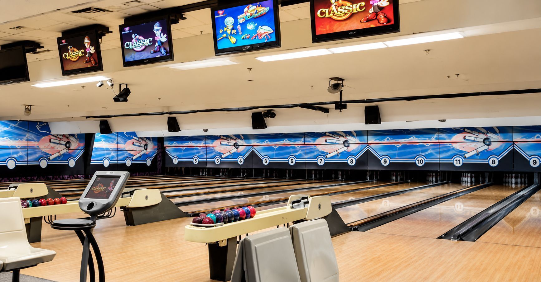 Empty bowling alley with glossy lanes, ball return, seating, and overhead score monitors.