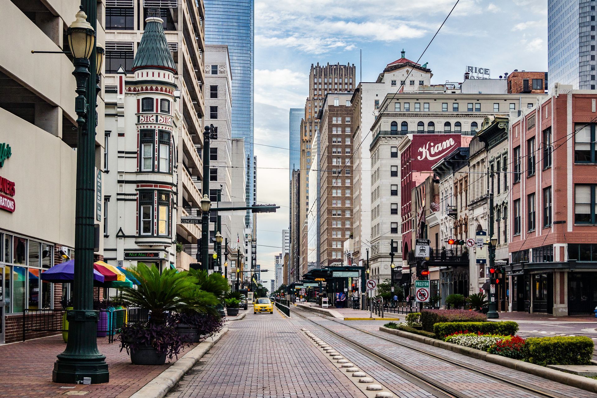 City street lined with buildings, trolley tracks down the center, cloudy sky.