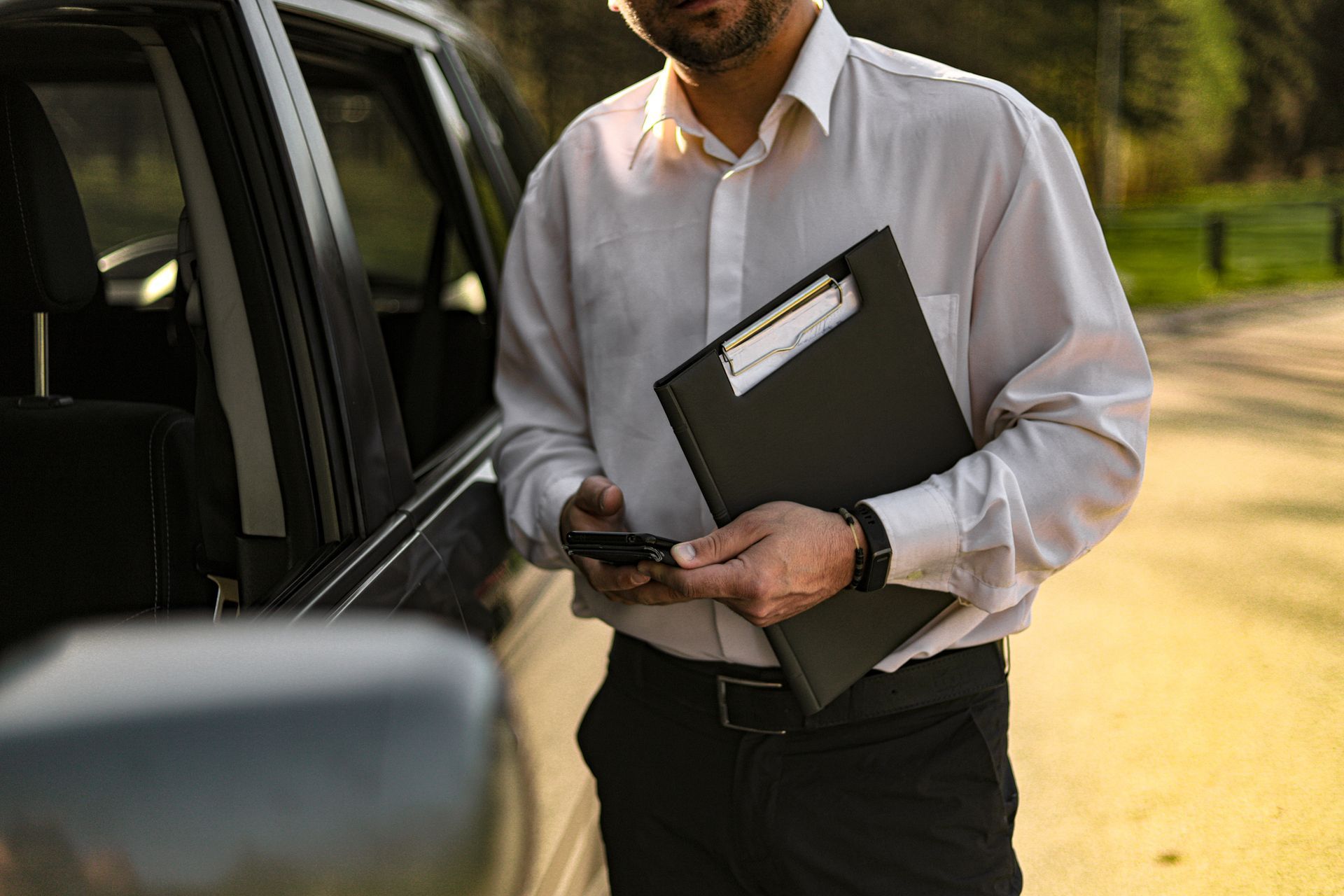 Man in white shirt near a car, holding a clipboard and phone. Outdoors. Man in white shirt near a car, holding a clipboard and phone. Outdoors.