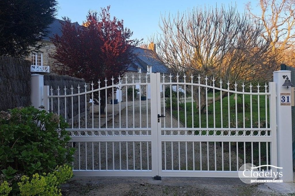 Portail d'allée en métal blanc devant une maison et un jardin, avec des arbres et des arbustes derrière.
