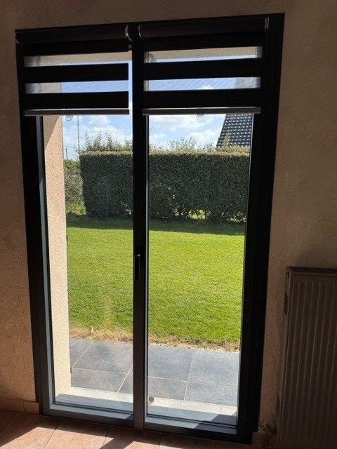 Porte-fenêtre coulissante avec stores, donnant sur un jardin ensoleillé avec pelouse et haie.