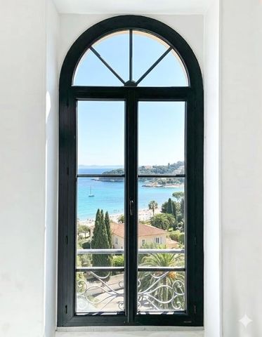 Une fenêtre cintrée noire encadre une vue lumineuse sur la mer, avec une eau bleue et le rivage au loin.