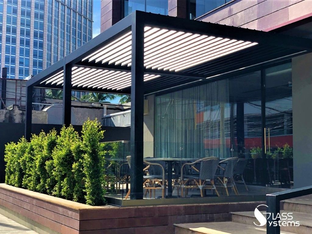 Terrasse extérieure moderne avec pergola noire, parois vitrées, tables et arbustes verts à côté d'un bâtiment.