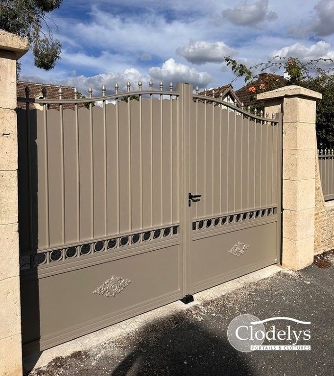 Portail d'allée en métal beige avec ornement décoratif, placé entre des piliers de pierre sous un ciel nuageux