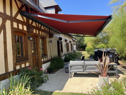 Terrasse attenante à une maison rustique en bois avec un auvent rouge, des sièges gris et des plantes en pot dans un jardin ensoleillé