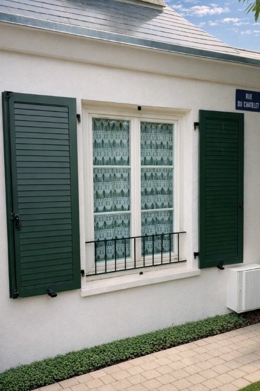 Mur d'une maison en stuc blanc avec une fenêtre, des volets verts, des rideaux à motifs et une allée pavée.