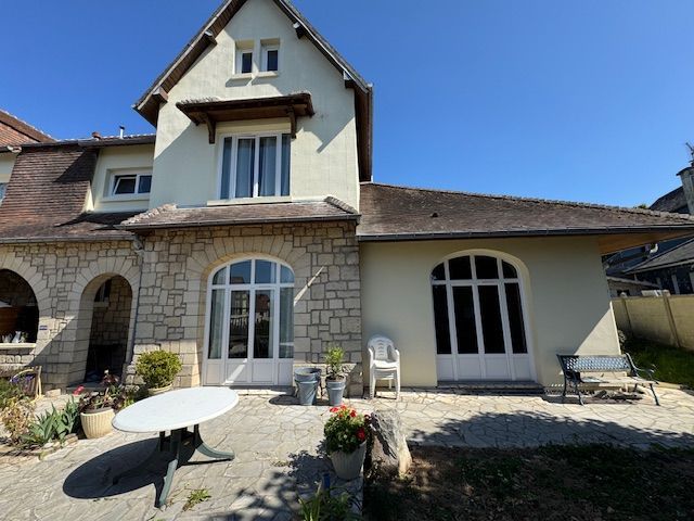 Patio d'une maison en pierre avec fenêtres cintrées, table blanche, fleurs en pot et ciel bleu