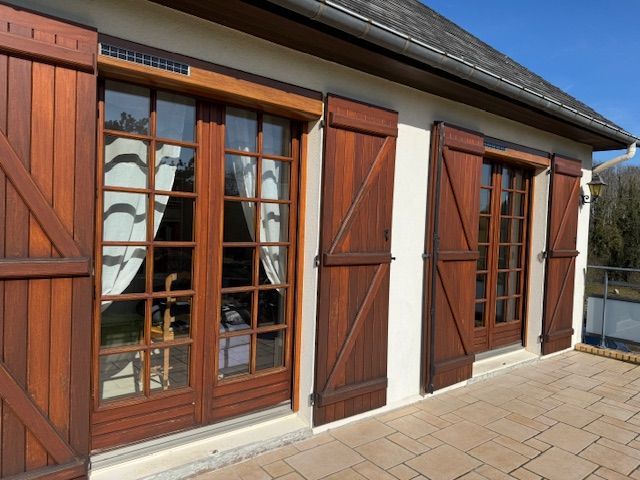 Portes-fenêtres et volets en bois donnant sur une façade de maison ensoleillée avec pavage en pierre.